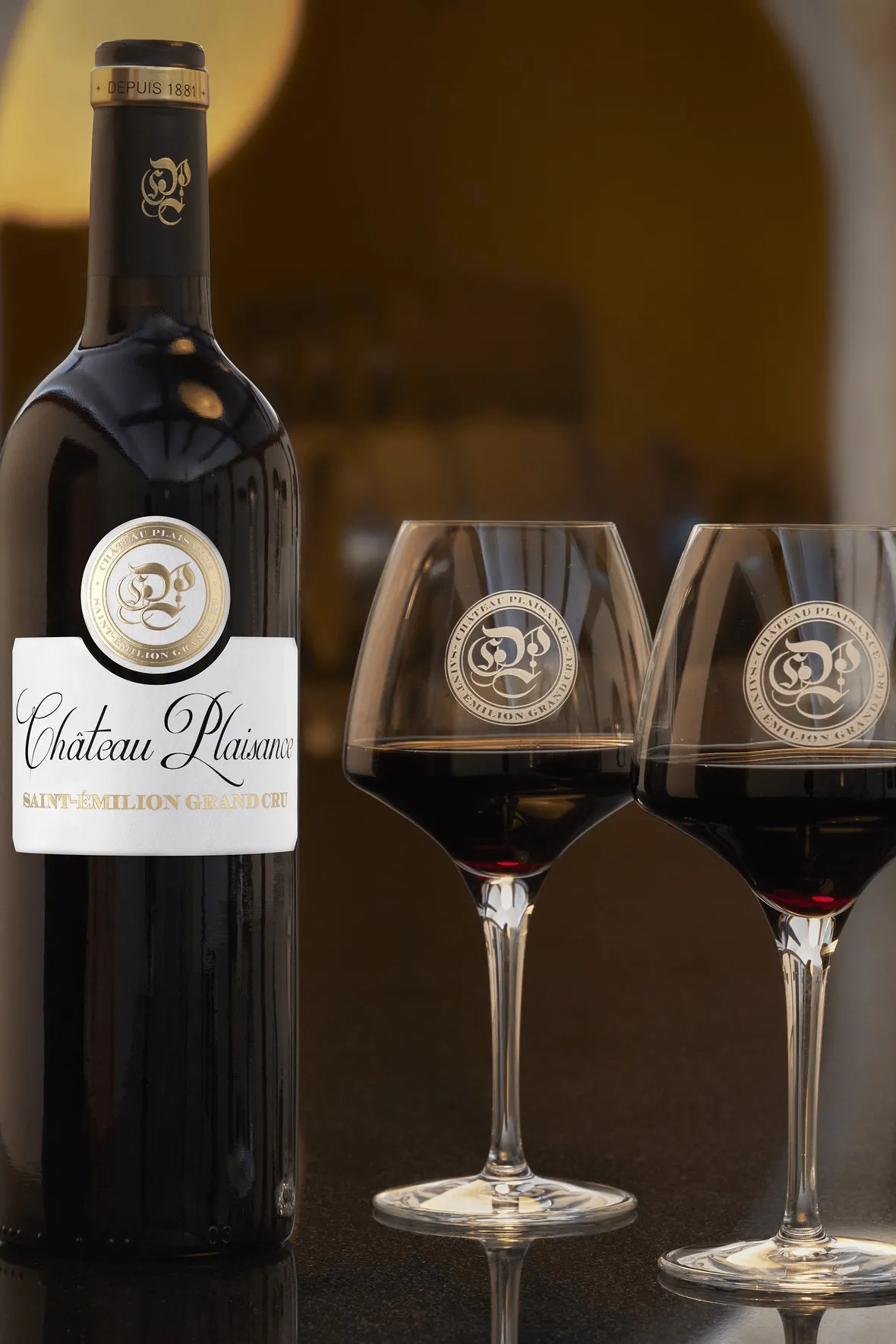 A photograph of a bottle of Château Plaisance award winning Saint-Émilion Grand Cru red wine with two glasses on the Château's tasting table.