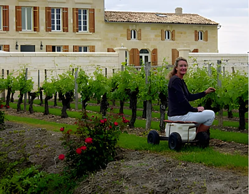 A woman working in the vineyard using a small utility cart while pruning vines, with the château building visible in the background.
