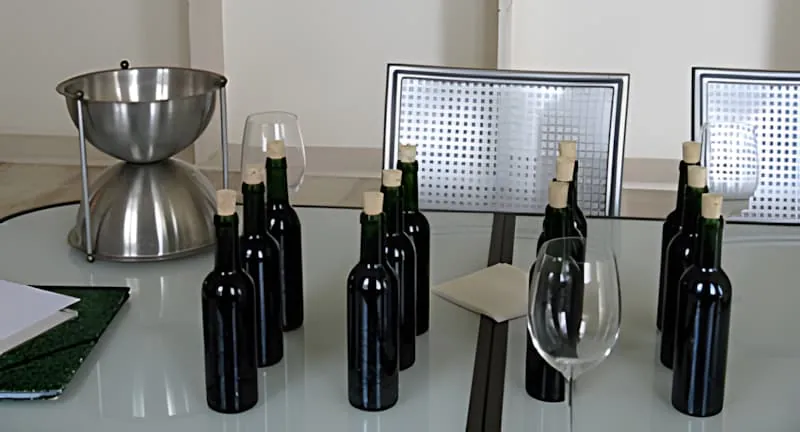 A formal wine tasting setup with multiple dark wine bottles, a silver spittoon, and wine glasses arranged on a table.