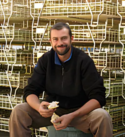 A smiling man in dark clothing sitting among stacked metal wine bottle crates during the bottling process.