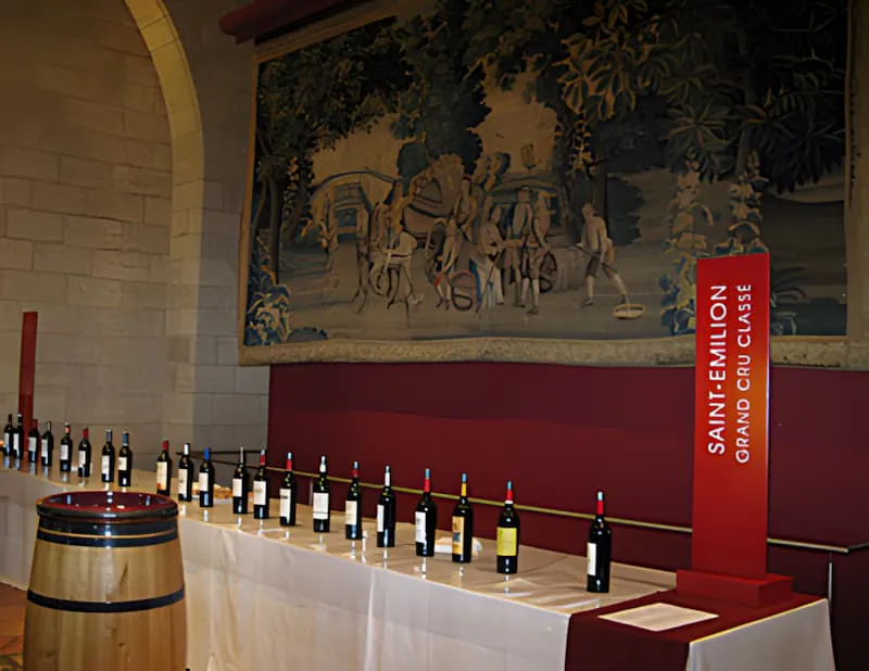 A long table lined with numerous bottles of red wine, set up for a professional tasting event in a room with a large tapestry.