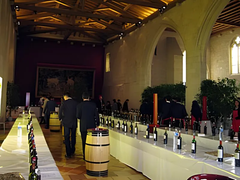 An elegant stone-vaulted hall with high ceilings where people are conducting wine tastings at tables lined with numerous bottles.