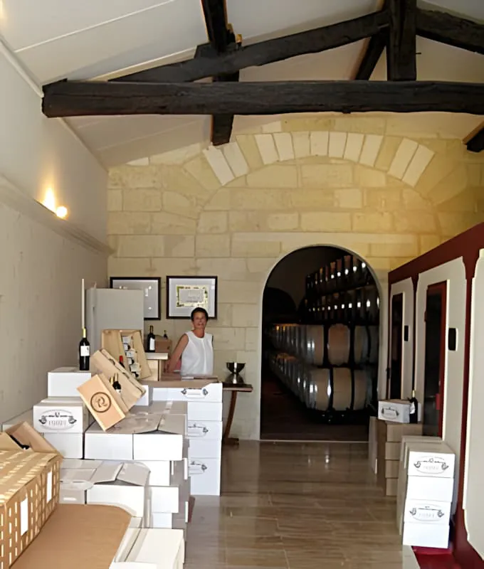 A woman standing behind a counter in a wine shop, surrounded by boxes of wine, with a view into the barrel room in the background.