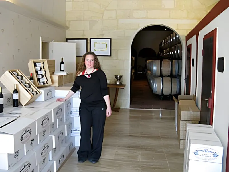 A young woman in dark clothing standing in a wine tasting room with boxes of wine and a view into the barrel cellar behind her.