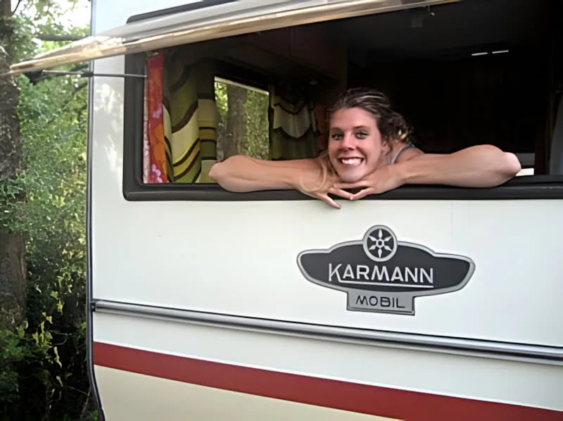 A smiling young woman with blonde hair leaning out of what appears to be a mobile home or trailer window.