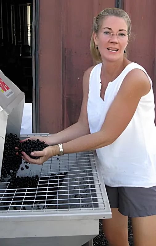 A blonde woman in a white tank top smiling while sorting dark grapes on a sorting table outside a building.