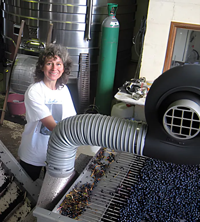 A woman with curly hair smiling while working at a grape sorting station surrounded by winemaking equipment in a cellar.