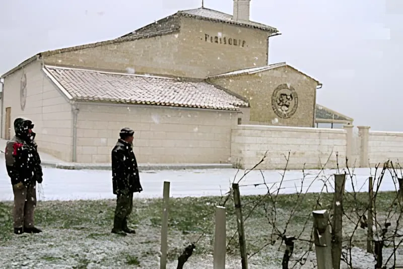Two workers in dark winter clothing standing near a stone winery building during heavy snowfall, with bare vineyard rows visible in the foreground.