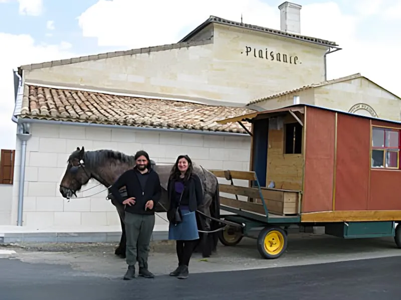 Two people standing next to a horse and colorful traveling wagon in front of a white building with