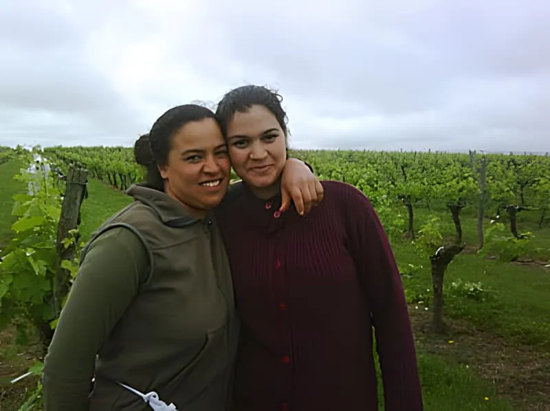 Two smiling women standing close together in a vineyard, one wearing a light colored jacket and the other in darker clothing.