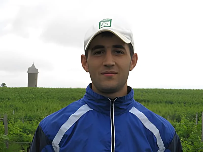 A smiling young man wearing a blue and white track suit jacket and cap, with a water tower visible in the green countryside background.