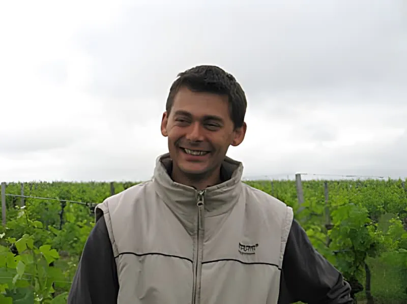 A cheerful young man in a light-colored jacket standing in front of lush green vineyard rows, smiling warmly at the camera.