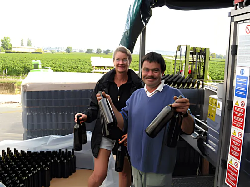A vineyard worker, smiling, is loading empty wine bottles into metal cages on a conveyor belt at Château Plaisance.